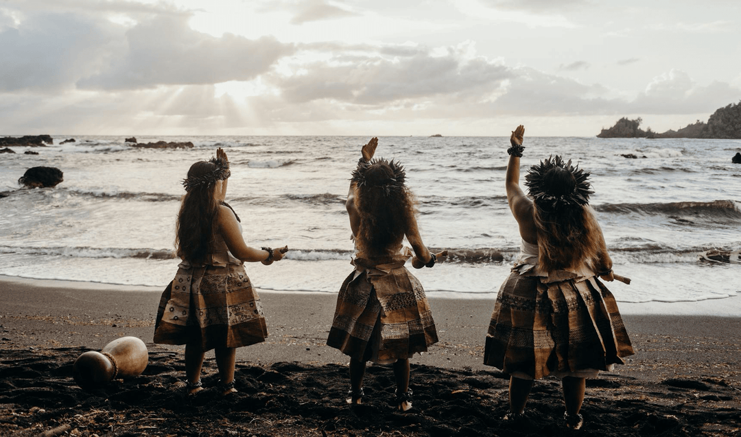 Three girls standing on a Hawaiian beach facing the ocean, jumping together – symbolizing Hawaiian culture and lifestyle.