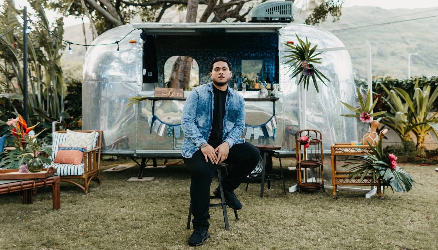 man sitting on a stool in an outdoor environment with a silver trailer in the background