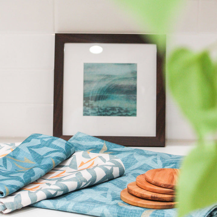 Lele Dining Placemat layred with Lele Napkins, and wooden coasters.  A framed Kilo Hōkū print graces the background.