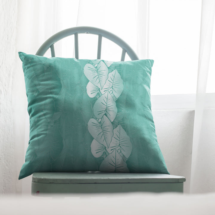 Teal pillow with white kalo leaf pattern on a light green chair against a white curtain background