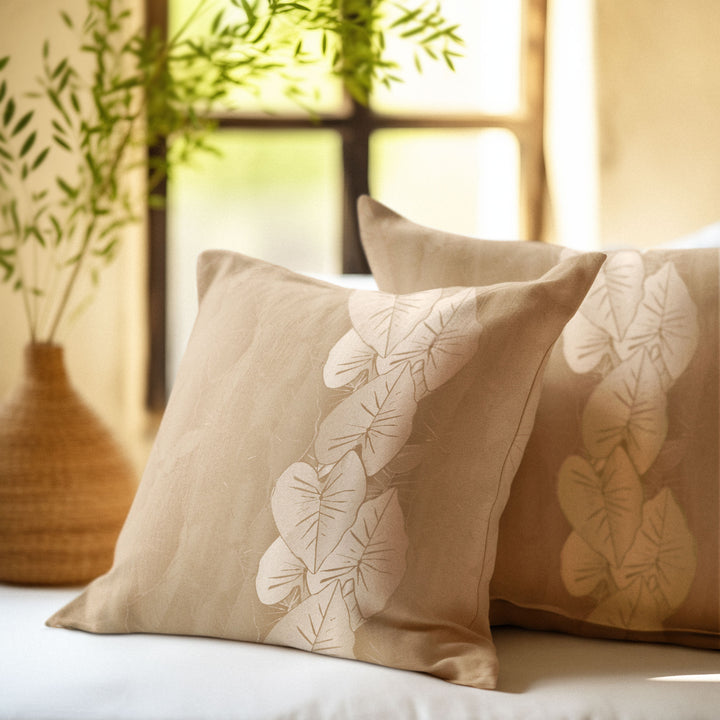 Beige pillows with kalo leaf patterns on a couch, with a blurred plant in the background.
