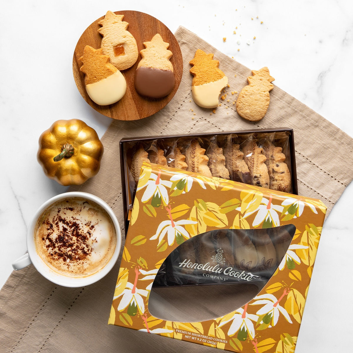A styled flat-lay of Honolulu Cookie Company cookies on a beige napkin over a white marble surface. The scene includes a box of assorted cookies, several acorn-shaped cookies on a wooden plate, a small gold pumpkin, and a white mug of coffee with cocoa powder.