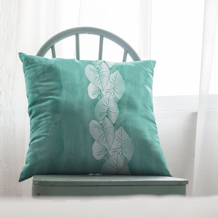 Teal pillow with white kalo leaf pattern on a light green chair against a white curtain background