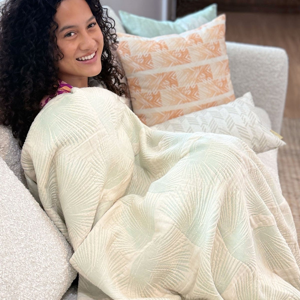 Woman sitting on a couch wrapped in a sage textured throw blanket with decorative pillows in the background.