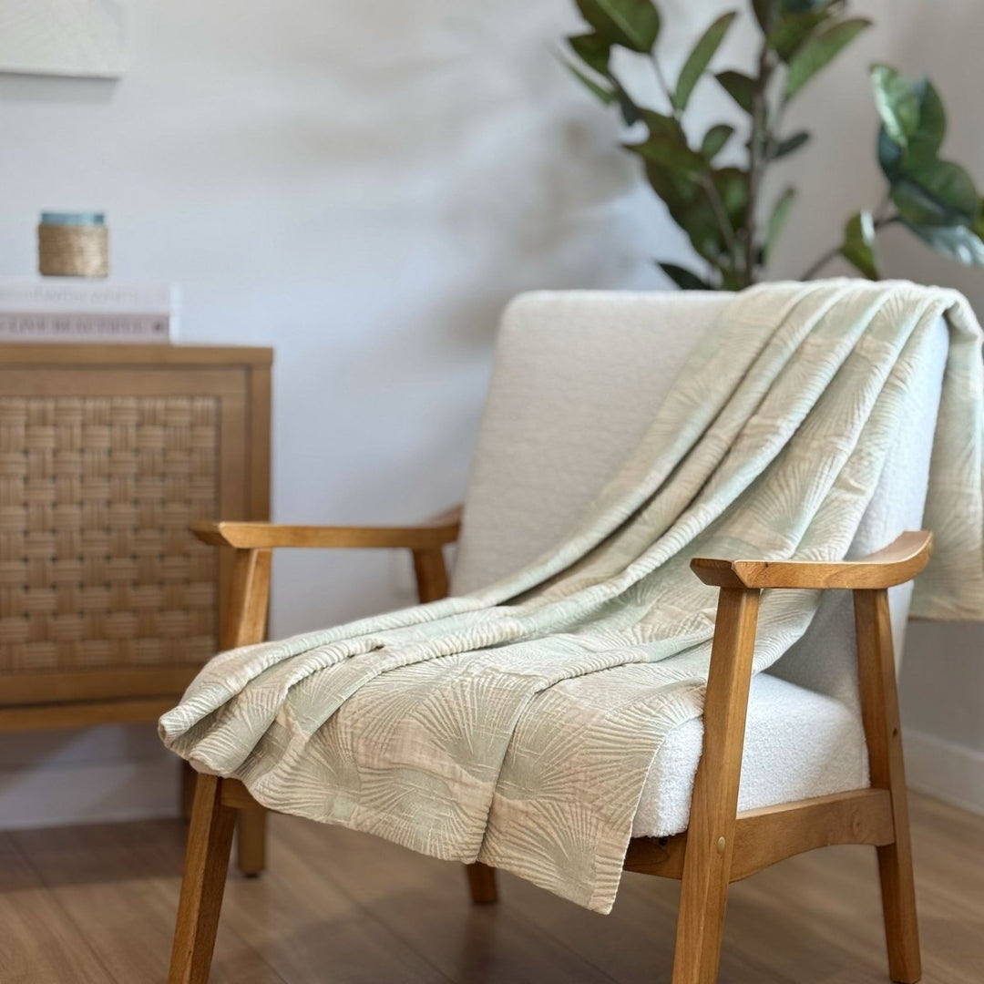Light green or sage textured throw blanket draped over a wooden armchair in a room with a plant and wooden side table.
