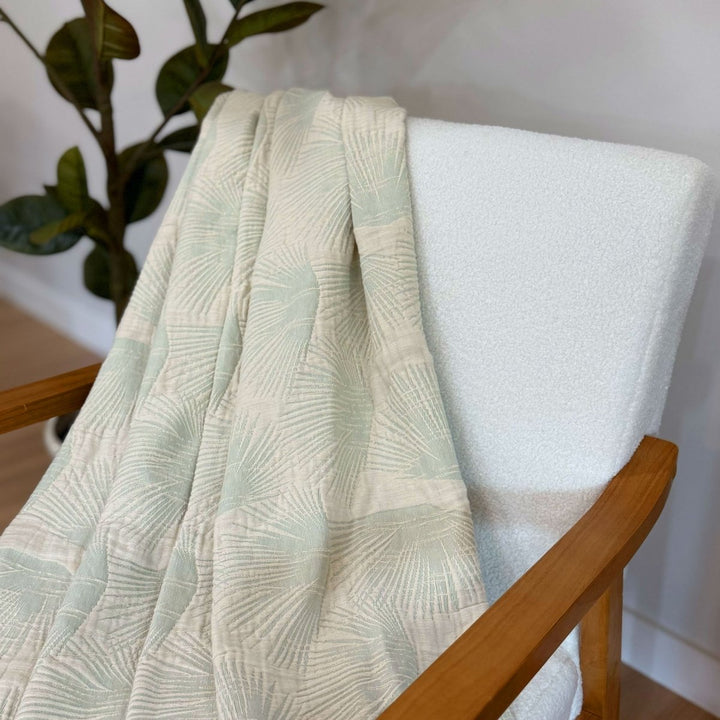 Light green or sage textured throw blanket draped over a white chair with a plant in the background
