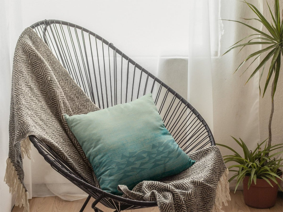 'Ōlali Square Pillowcase resting on a throw on a comfortable chair in a corner of cozy area with filtered light a small tree