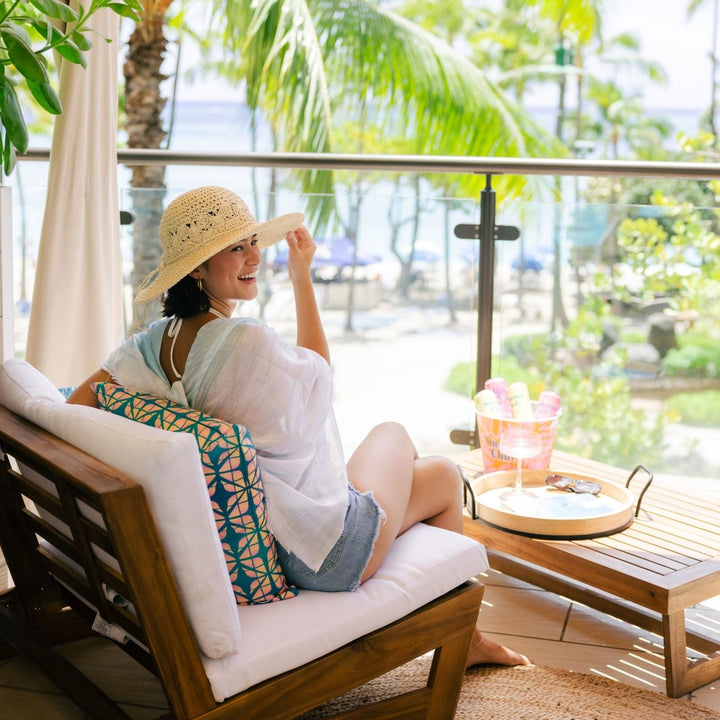 Woman sitting on a wooden chair with white cushions, wearing a straw hat and white shirt, in a tropical setting.