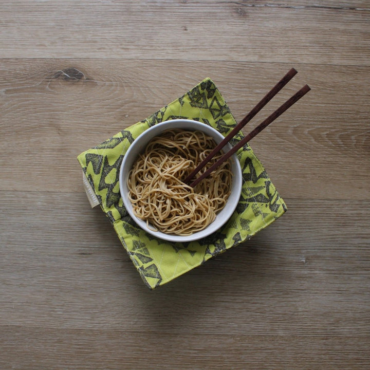 Bowl of noodles with chopsticks on a green patterned bowl cozy on a wooden surface