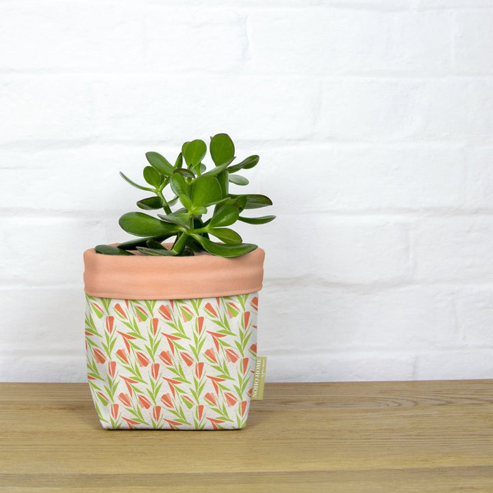 A green succulent is planted in a fabric planter with a peach and green floral pattern. The planter has a solid peach cuff and is placed on a wooden surface against a white brick wall.