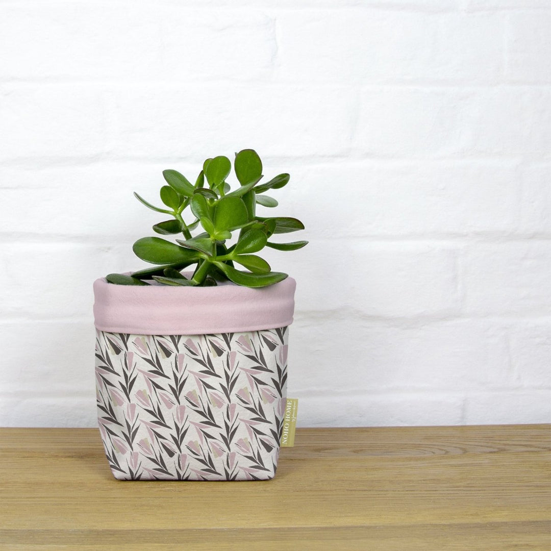 Rose Lokelani Fabric Planter - A green succulent is planted in a fabric planter with a pink and gray floral pattern. The planter has a solid baby pink cuff and is placed on a wooden surface against a white brick wall.