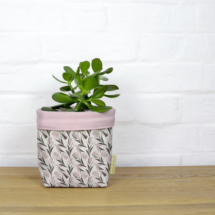 Rose Lokelani Fabric Planter - A green succulent is planted in a fabric planter with a pink and gray floral pattern. The planter has a solid baby pink cuff and is placed on a wooden surface against a white brick wall.