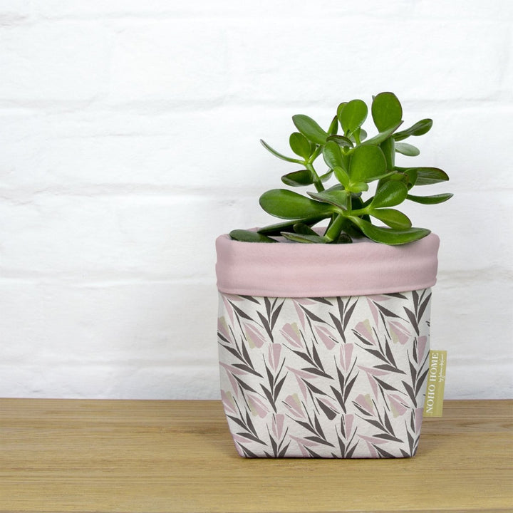 Rose Lokelani Fabric Planter - Medium - A green succulent is planted in a fabric planter with a pink and gray floral pattern. The planter has a solid baby pink cuff and is placed on a wooden surface against a white brick wall.