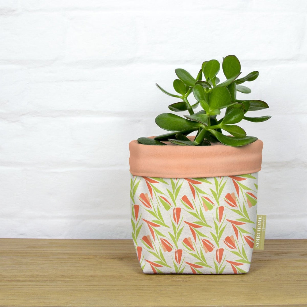 A green succulent is planted in a fabric planter with a peach and green floral pattern. The planter has a solid peach cuff and is placed on a wooden surface against a white brick wall.