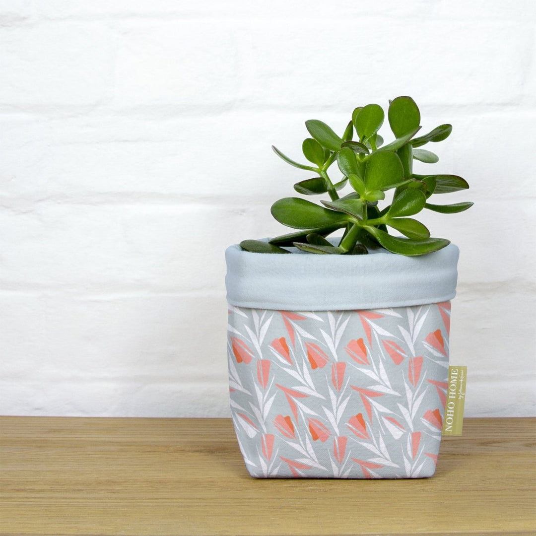 A green succulent is planted in a fabric planter with a light blue and peach floral pattern. The planter has a solid baby blue cuff and is placed on a wooden surface against a white brick wall.