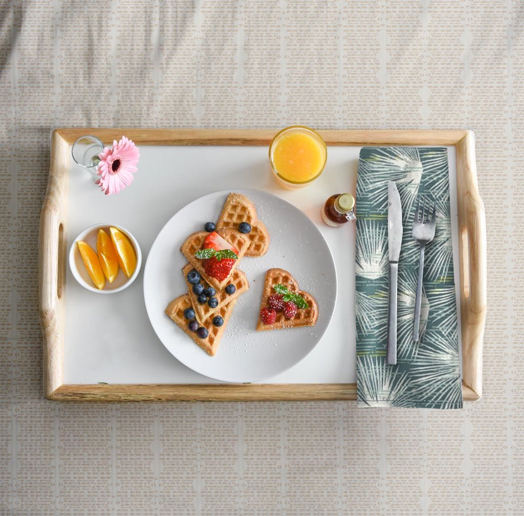 Breakfast tray  with a waffle plate, orange wedges in a bowl, as well as pink flower in vase, orange juice and small jar of syrup.  Folded Light Blue and Beige Loulu napkin with silverware resting on top completes the look.