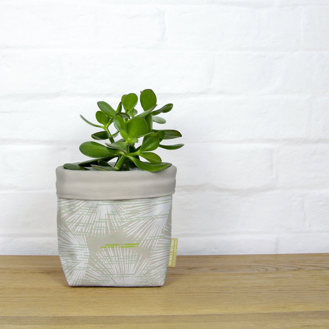 A green succulent is planted in a fabric planter with a beige and ivory palm leaf design. The planter has a solid beige cuff and is placed on a wooden surface against a white brick wall.