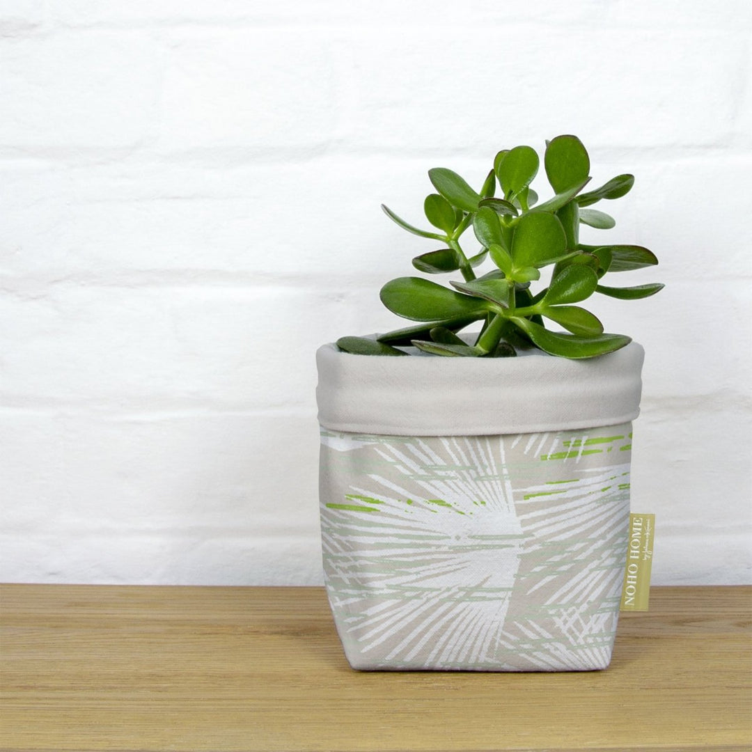 A green succulent is planted in a fabric planter with a beige and ivory palm leaf design. The planter has a solid beige cuff and is placed on a wooden surface against a white brick wall.