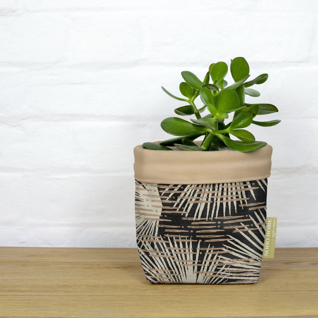 A green succulent is planted in a fabric planter with a taupe and black palm leaf design. The planter has a solid taupe cuff and is placed on a wooden surface against a white brick wall.