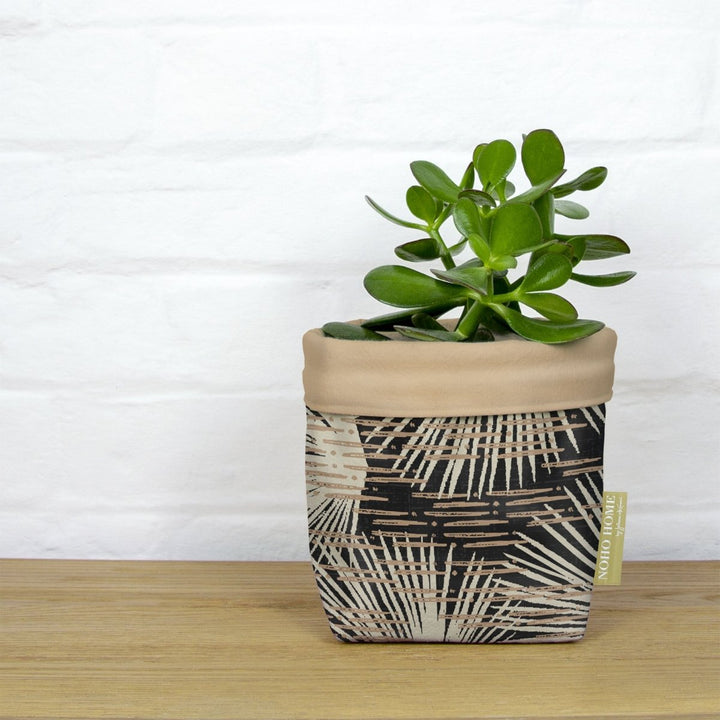 A green succulent is planted in a fabric planter with a taupe and black palm leaf design. The planter has a solid taupe cuff and is placed on a wooden surface against a white brick wall.