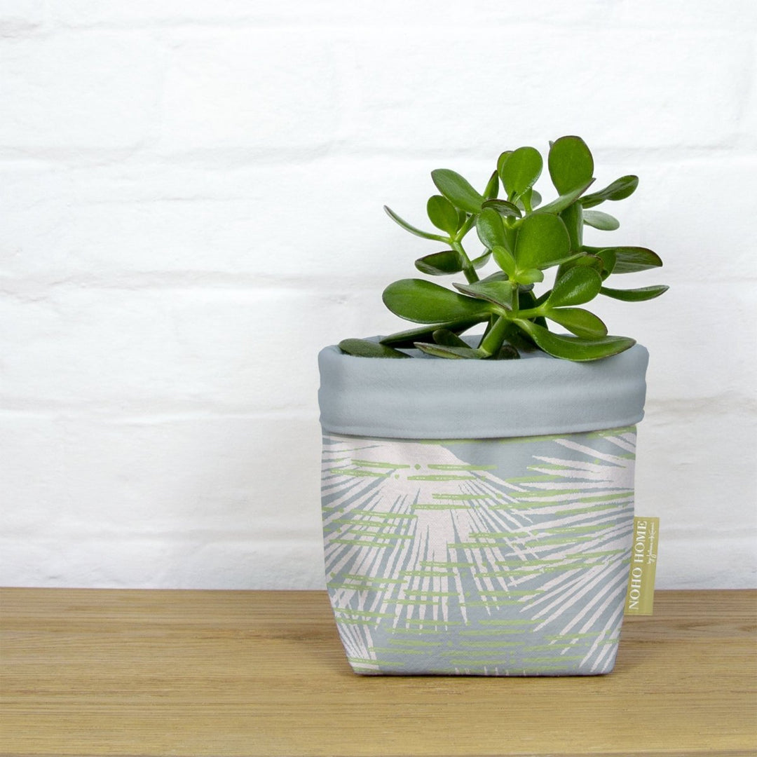A green succulent is planted in a fabric planter with a celery and light blue palm leaf design. The planter has a solid light blue cuff and is placed on a wooden surface against a white brick wall.