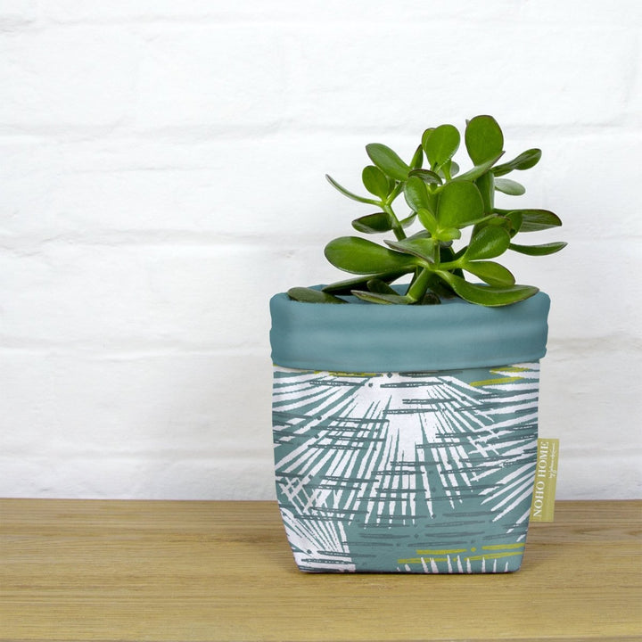 A green succulent is planted in a fabric planter with an ivory and teal palm leaf design. The planter has a solid teal cuff and is placed on a wooden surface against a white brick wall.