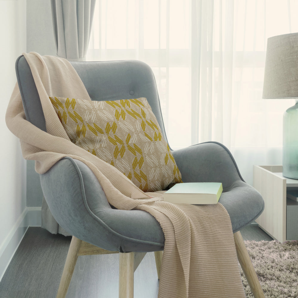 Peʻa Square Pillow propped on a light throw on a modern blue chair in a quiet space with filtered lighting and lamp on a table