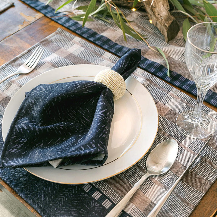 Black Premium Batik Dining Napkin Set with napkin ring resting on white ceramic plate on Papa Mu Placemat, glass challis and silverware.
